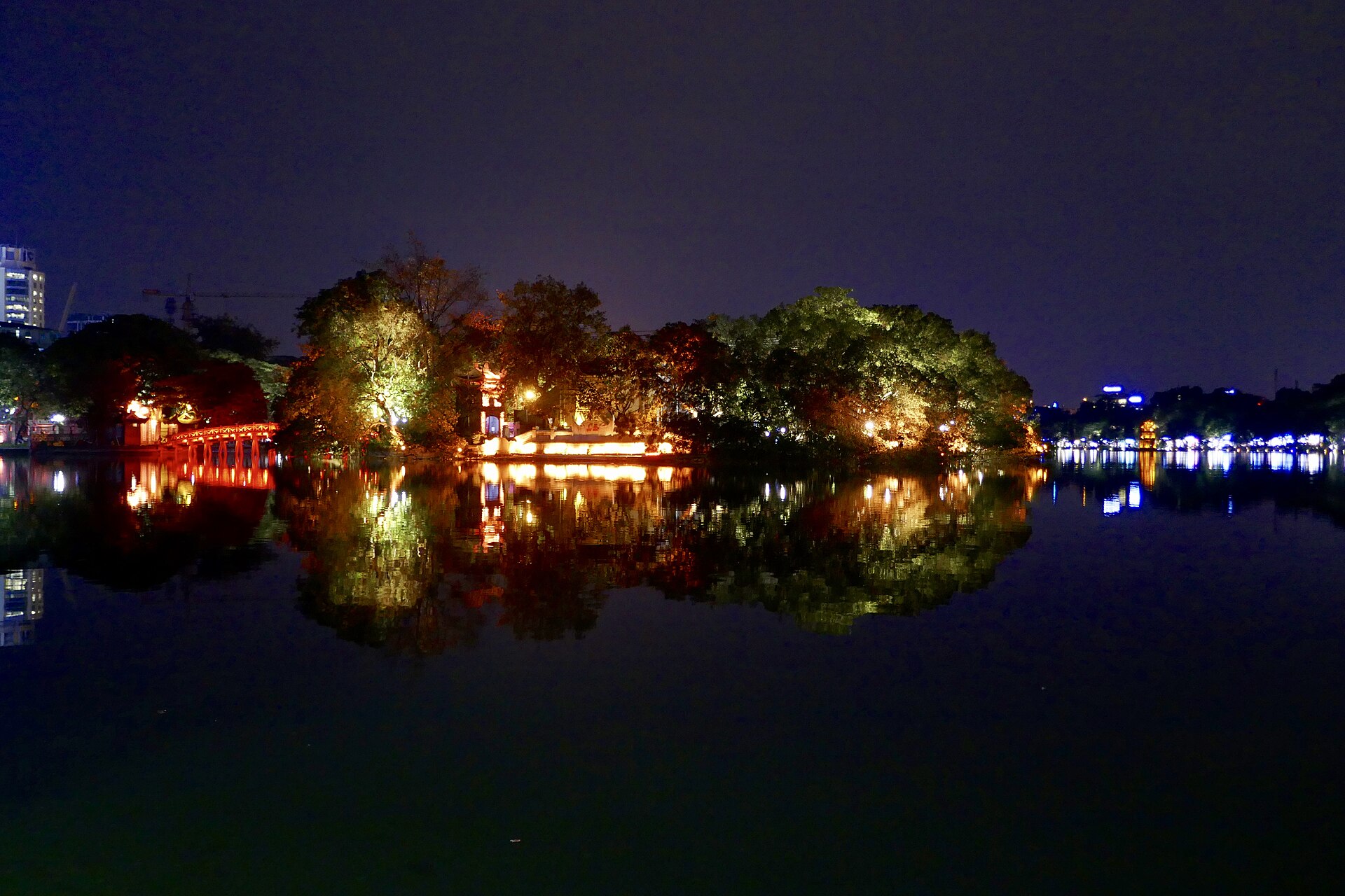 Lights from Thê Húc Bridge and Ngọc Sơn’s tree-covered island reflected in Hoàn Kiếm Lake at night.