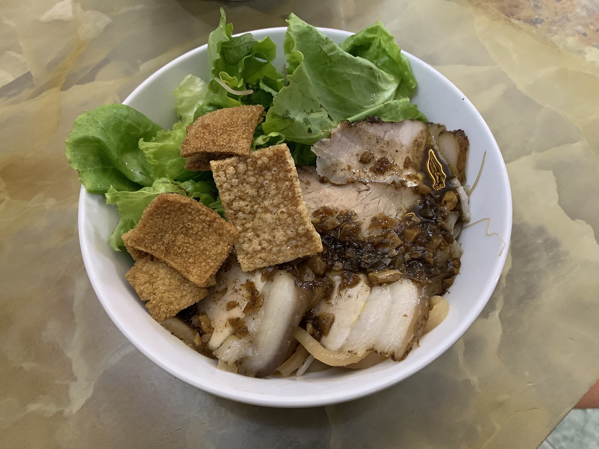 A bowl of cao lầu noodles topped with pork, fresh greens and crisp crackers.