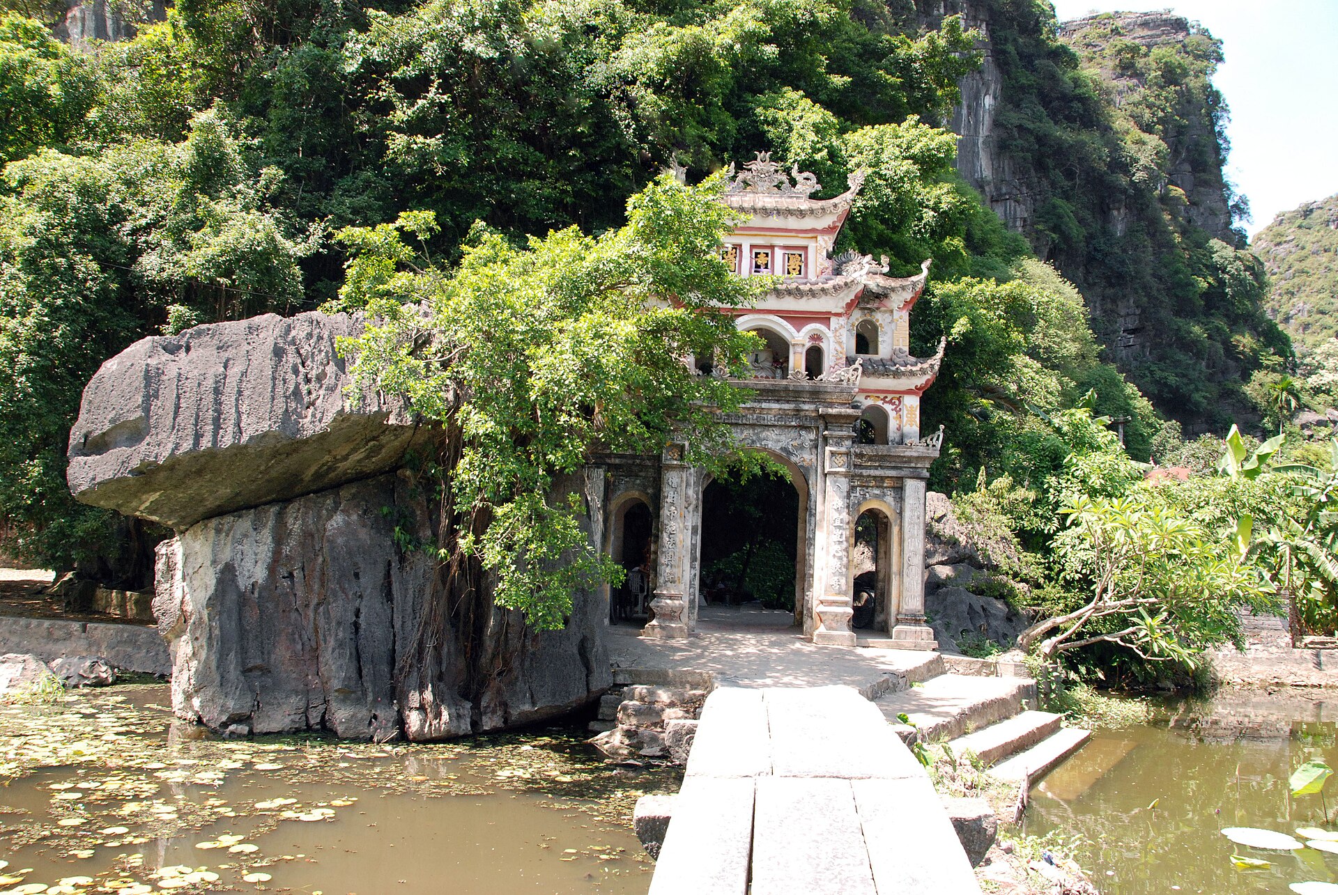 A stone bridge leads across water to Bích Động’s ornate gateway beneath a forested limestone cliff.