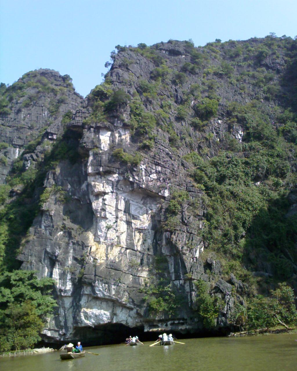 A small rowboat passes a tall limestone cliff on the water at Tam Cốc.